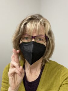 Author, voice actor Lisa Leonard, shown in photo wearing a black face mask. She has shoulder length blonde hair and purple and green plastic framed eye glasses. She is crossing her fingers, hoping she did not catch COVID during her recent conference attendance 