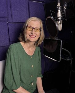 blonde woman wearing green shirt sits in VoiceOver booth near microphone and smiles at camera